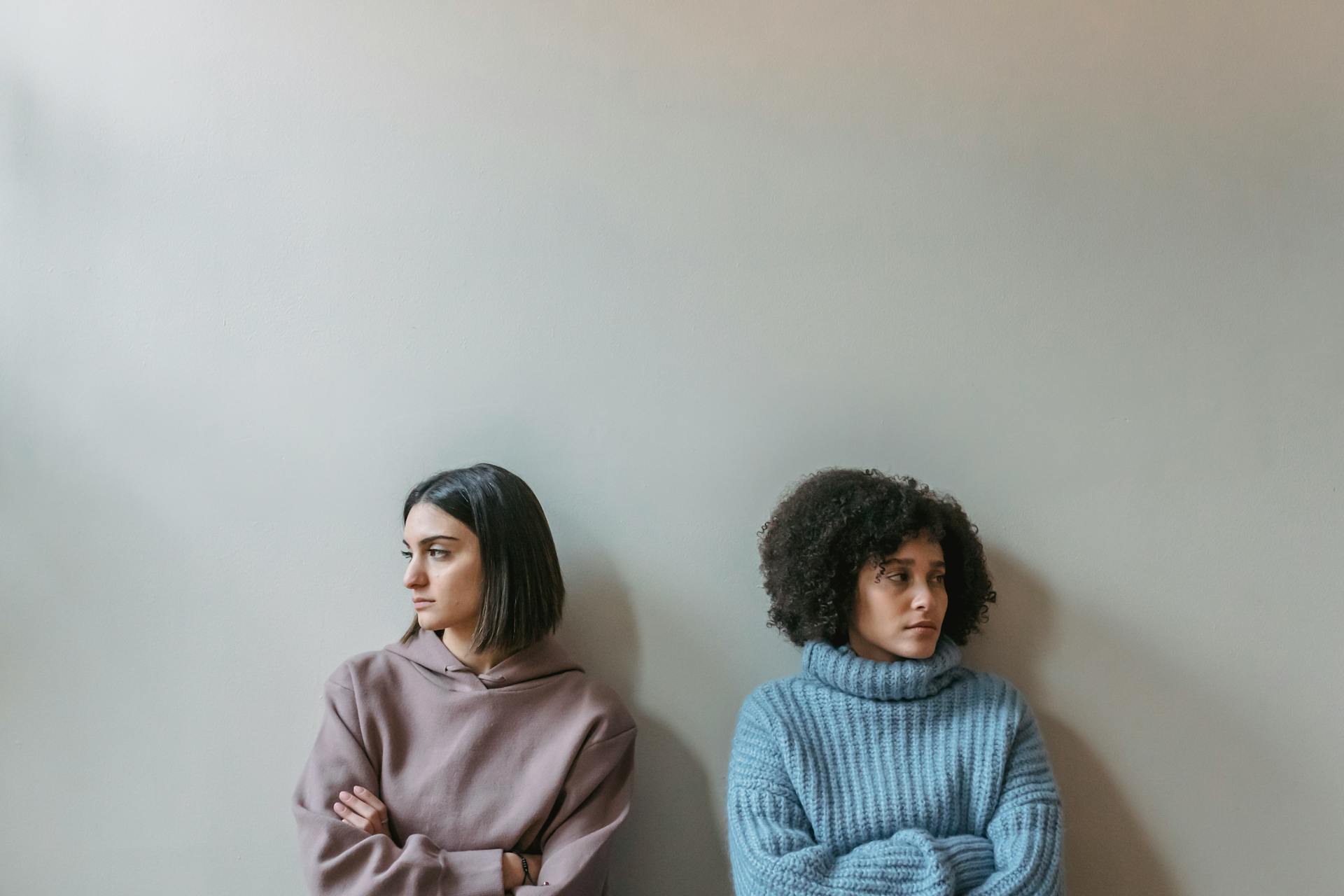 Sad multiracial female friends with crossed arms looking away and standing near white wall while having conflict in light room at home