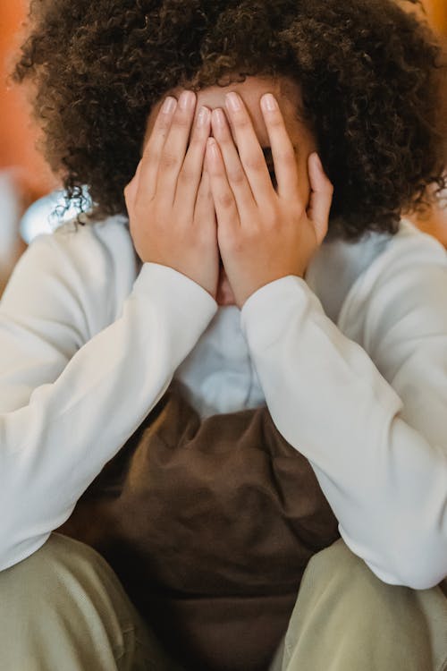 Unrecognizable sad ethnic female with curly hair in casual wear covering face while sitting with pillow in room at home