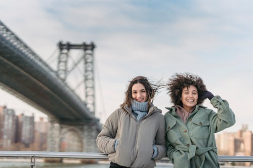 Delighted multiracial female friends in outerwear looking at camera while standing on embankment near bridge against cityscape on blurred background