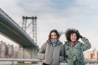 Delighted multiracial female friends in outerwear looking at camera while standing on embankment near bridge against cityscape on blurred background