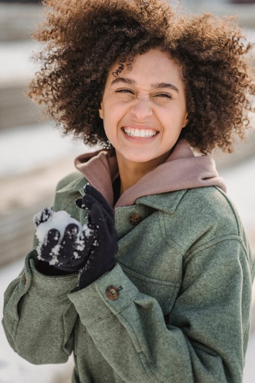 Joyful ethnic female in warm clothes and gloves looking away while playing snowballs on snowy street on blurred background in winter day