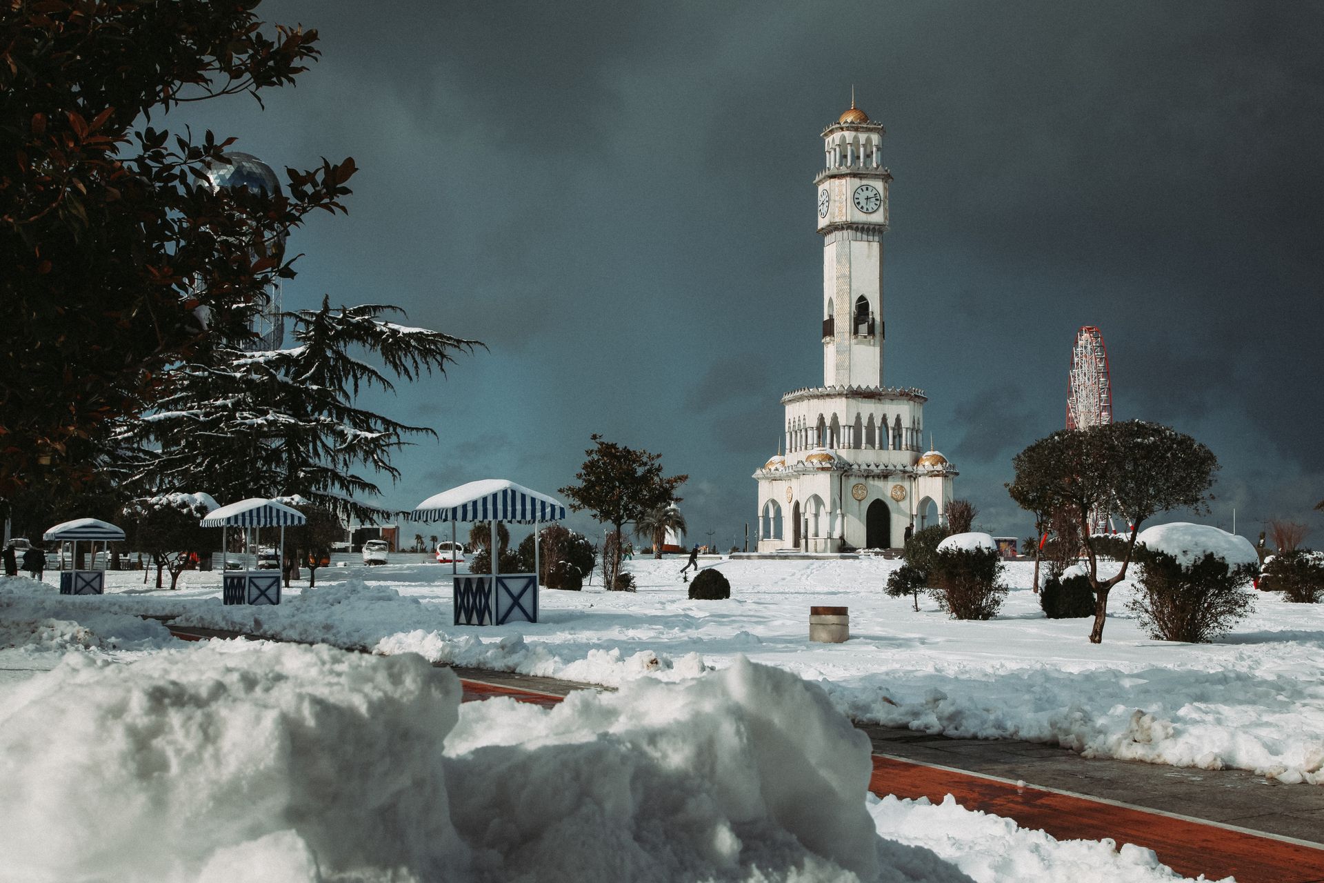Tall white clock tower on snowy terrain near wooden structures and green trees located against overcast sky in Georgia in Batumi city in winter time