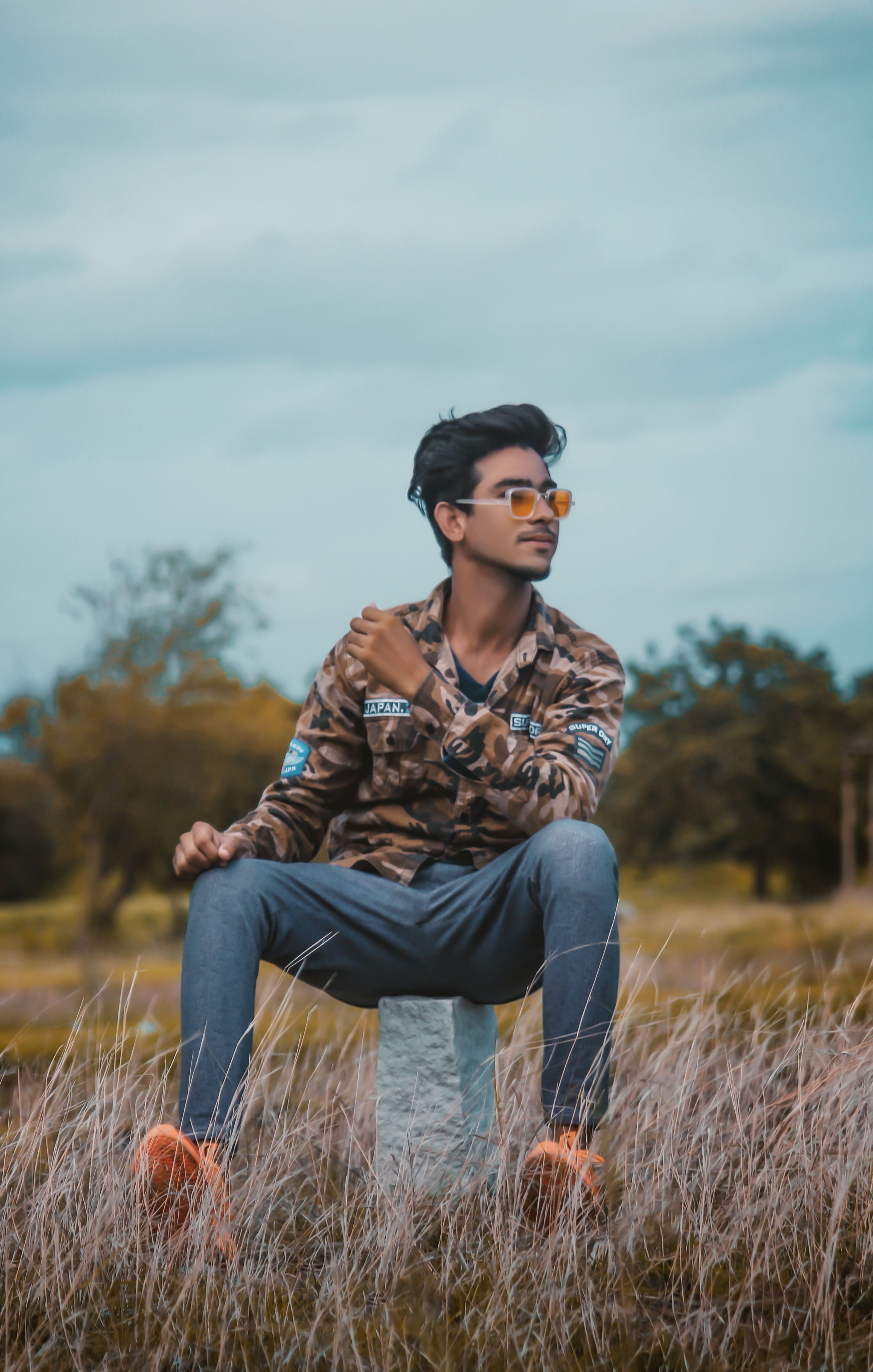 Casual young man in sunglasses sitting on rock in grassy field outdoors.
