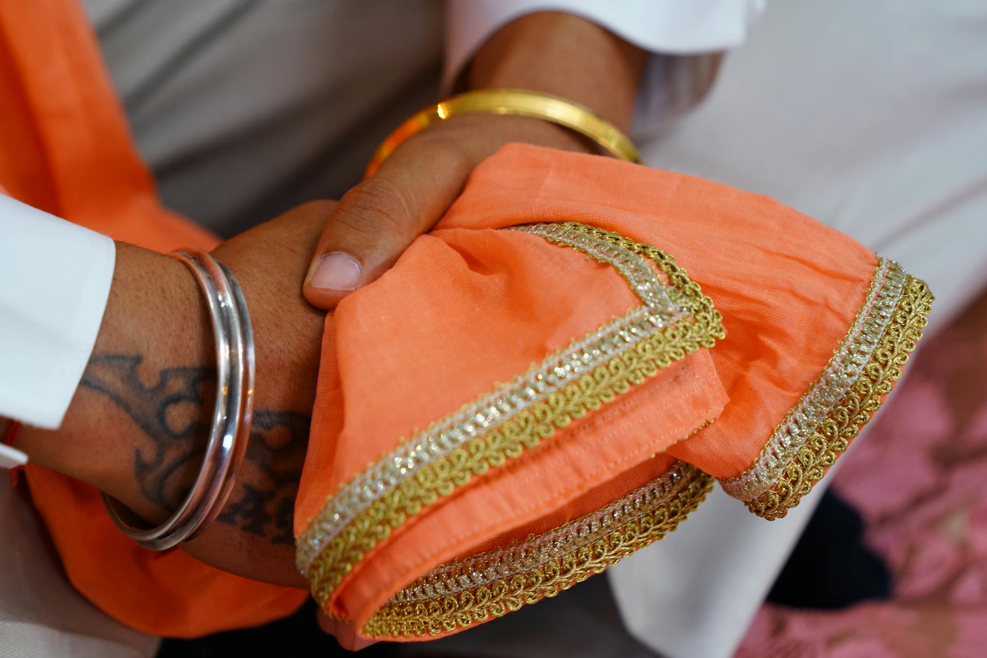 Crop unrecognizable Indian groom wearing traditional white wedding clothes and pink scarf with embroidery sitting with hands clasped