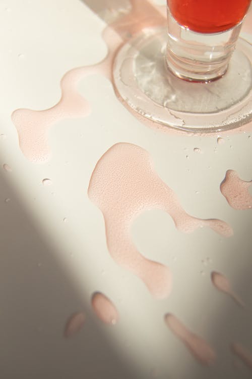 From above glass of red colored beverage spilled on white table in sunlight