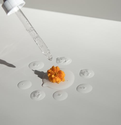 From above of drops of transparent moisturizing cosmetic product dripped by pipette and small fresh flower head placed on white table
