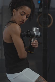 A woman in a gym uses a percussion massager on her arm, promoting muscle recovery and fitness.