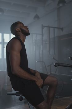 A fit African American man sits on gym equipment, highlighting a moment of rest during a workout.