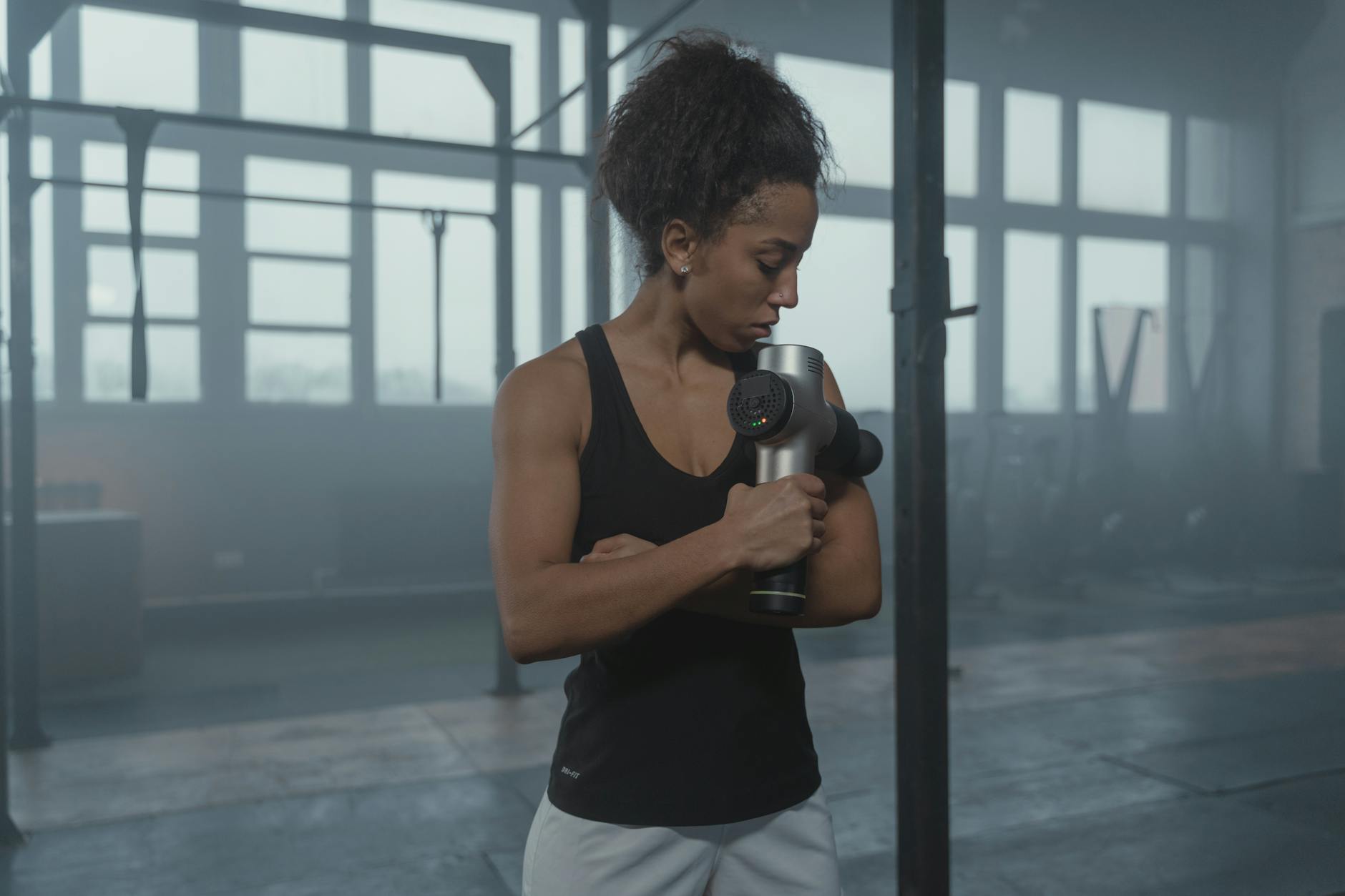 Fit woman using a massage gun in a well-lit gym studio for muscle recovery.