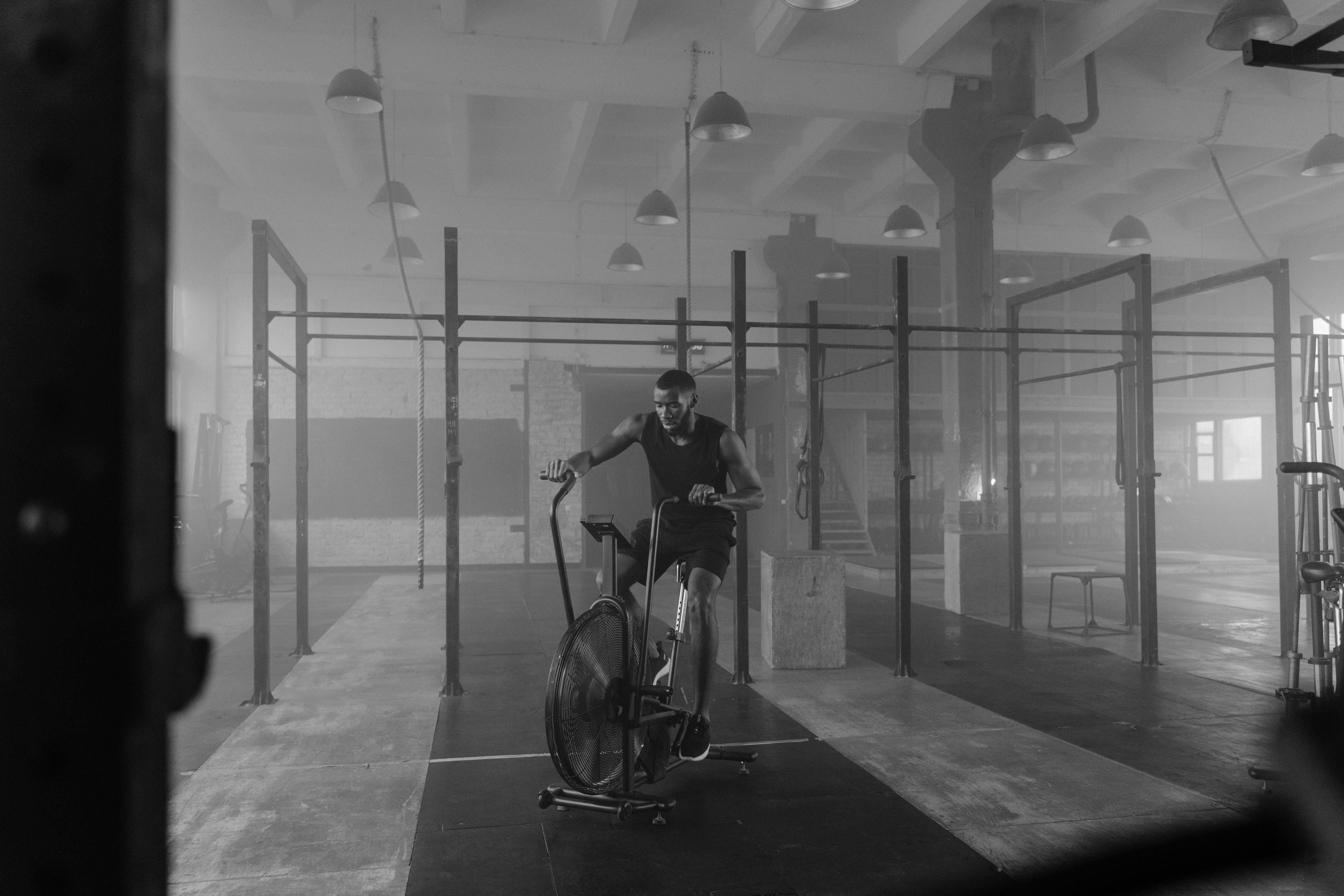 Man training on an air bike in a dimly lit gym, captured in black and white.