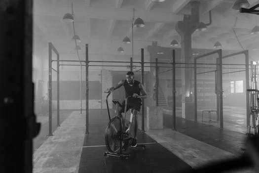 Man training on an air bike in a dimly lit gym, captured in black and white.