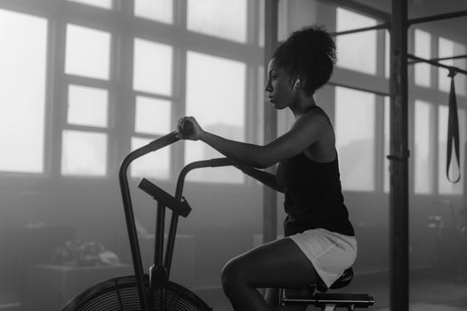 Black and white image of a woman working out on an air bike in a gym setting.