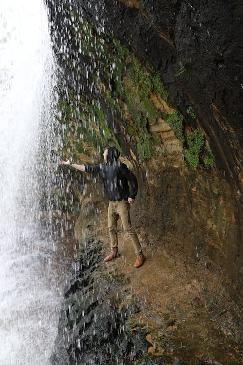 Side view traveler touching water drops while standing on ledge near scenery waterfall