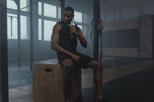 Free Fit man using a massage gun in a gym, wearing a tank top while seated on a wooden box. Stock Photo