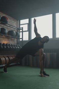 An athletic man performs a stretching exercise in a gym surrounded by fitness equipment.