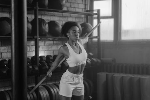 A woman in activewear stretching with a barbell in a monochrome gym setting.