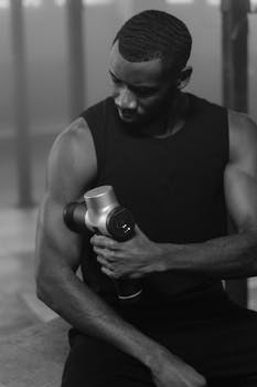 Muscular man in tank top using a massage gun for recovery in a black and white photo.