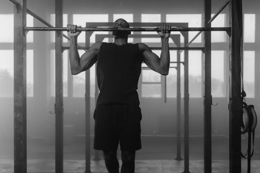 Black and white photo of a man performing pull-ups in an indoor gym setting, showcasing strength and fitness.