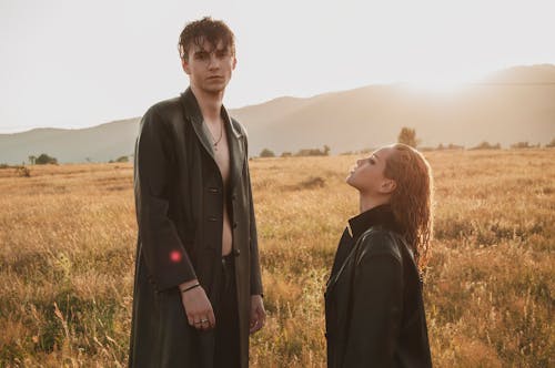 Extravagant couple wearing black jackets standing in meadow with dry grass against hills in sunny weather