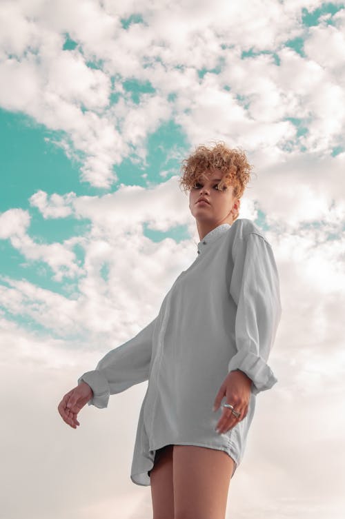 From below of eccentric female with stylish curly hairstyle standing against clouds floating over blue sky and looking away