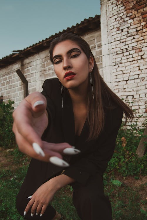 Free Serious female wearing black suit with trendy makeup standing against brick building and reaching hand to camera Stock Photo