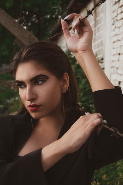 Provocative female with stylish makeup in black jacket standing near metal fence and looking away