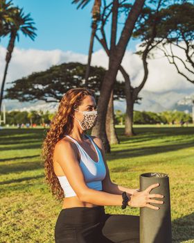 Woman in activewear with yoga mat kneeling in a sunny park with trees in the background.