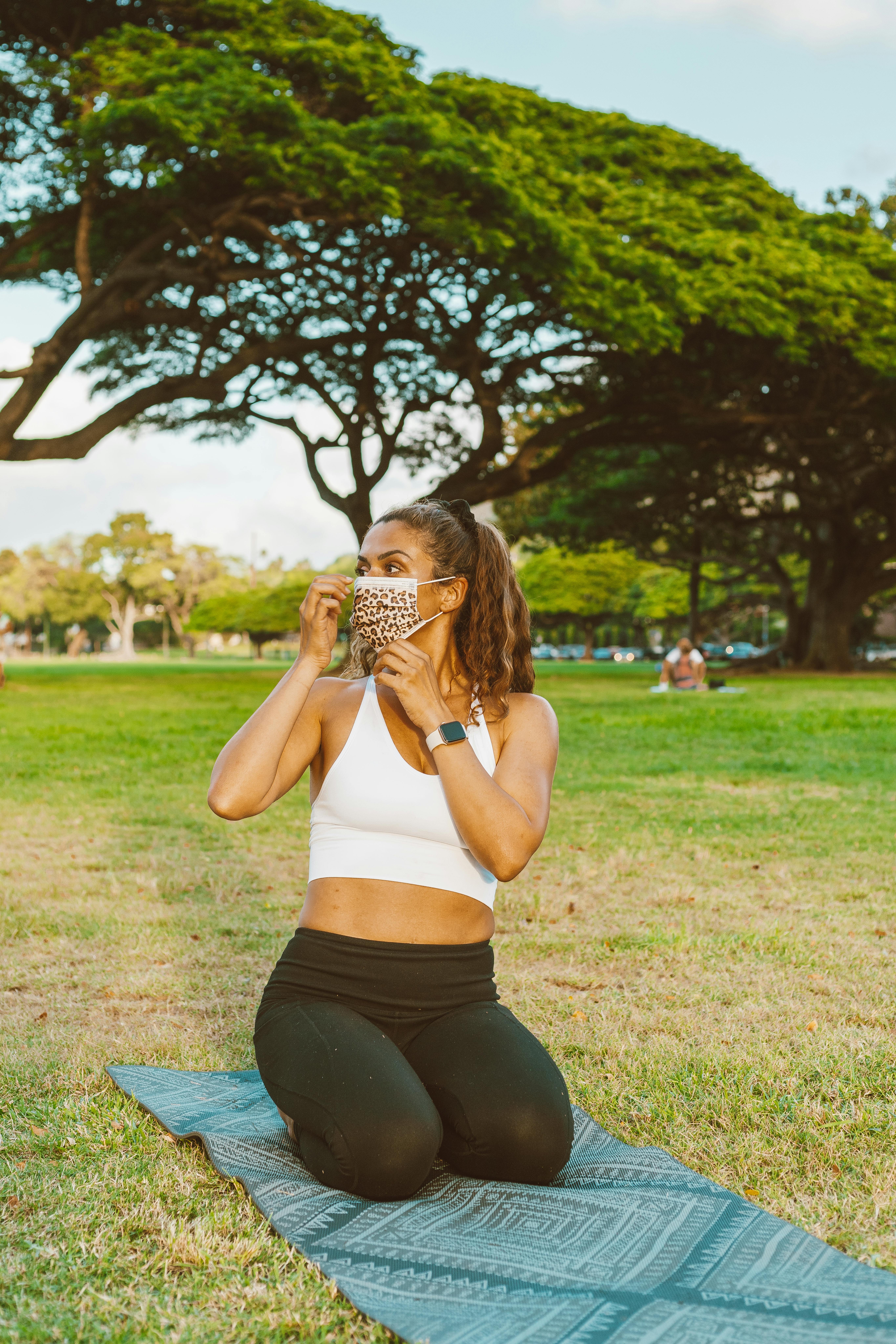 Woman kneeling on yoga mat in park, adjusting face mask, blending fitness with safety.