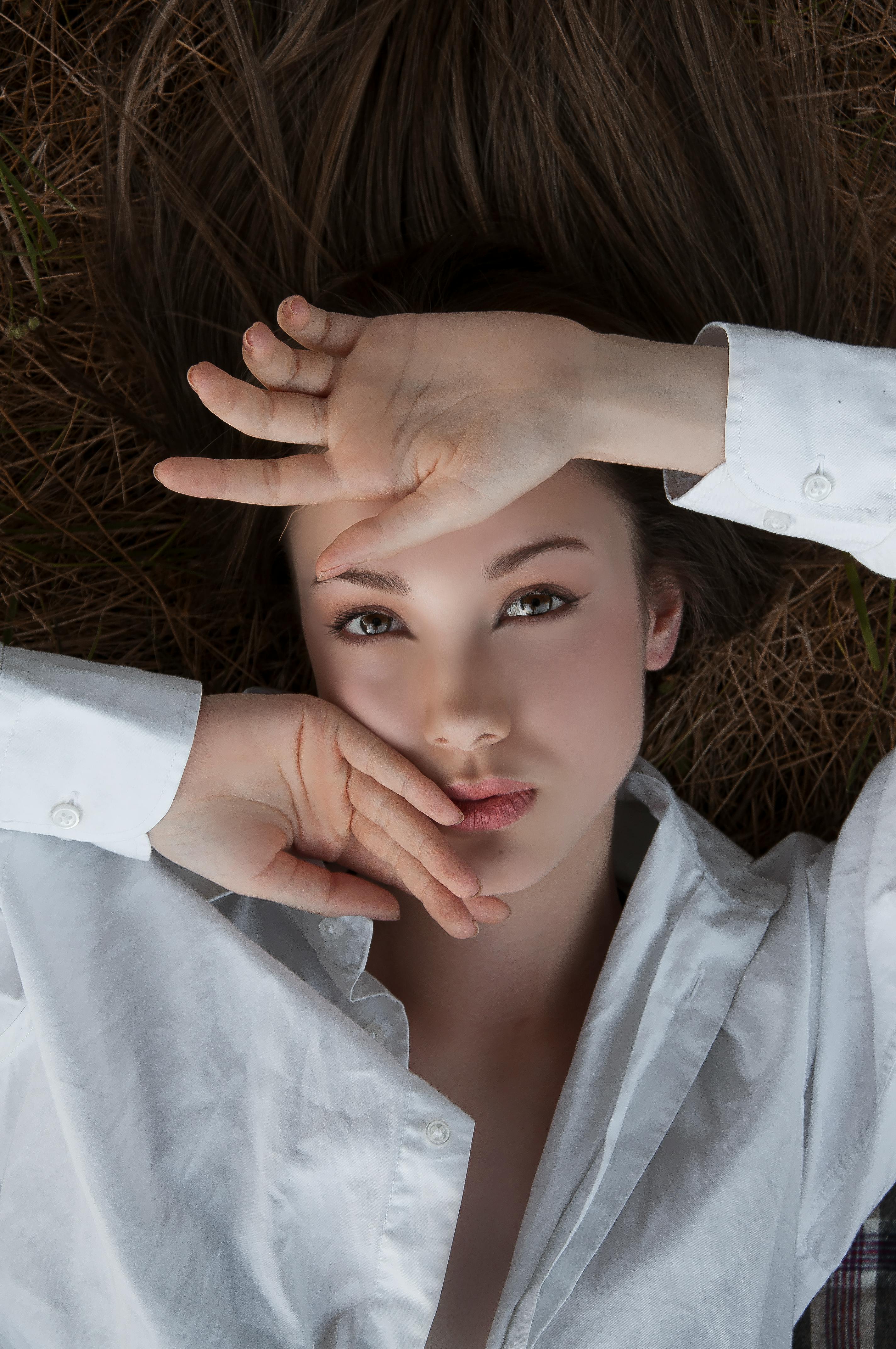 From above of alluring female with long hair touching face while lying on grass and looking at camera