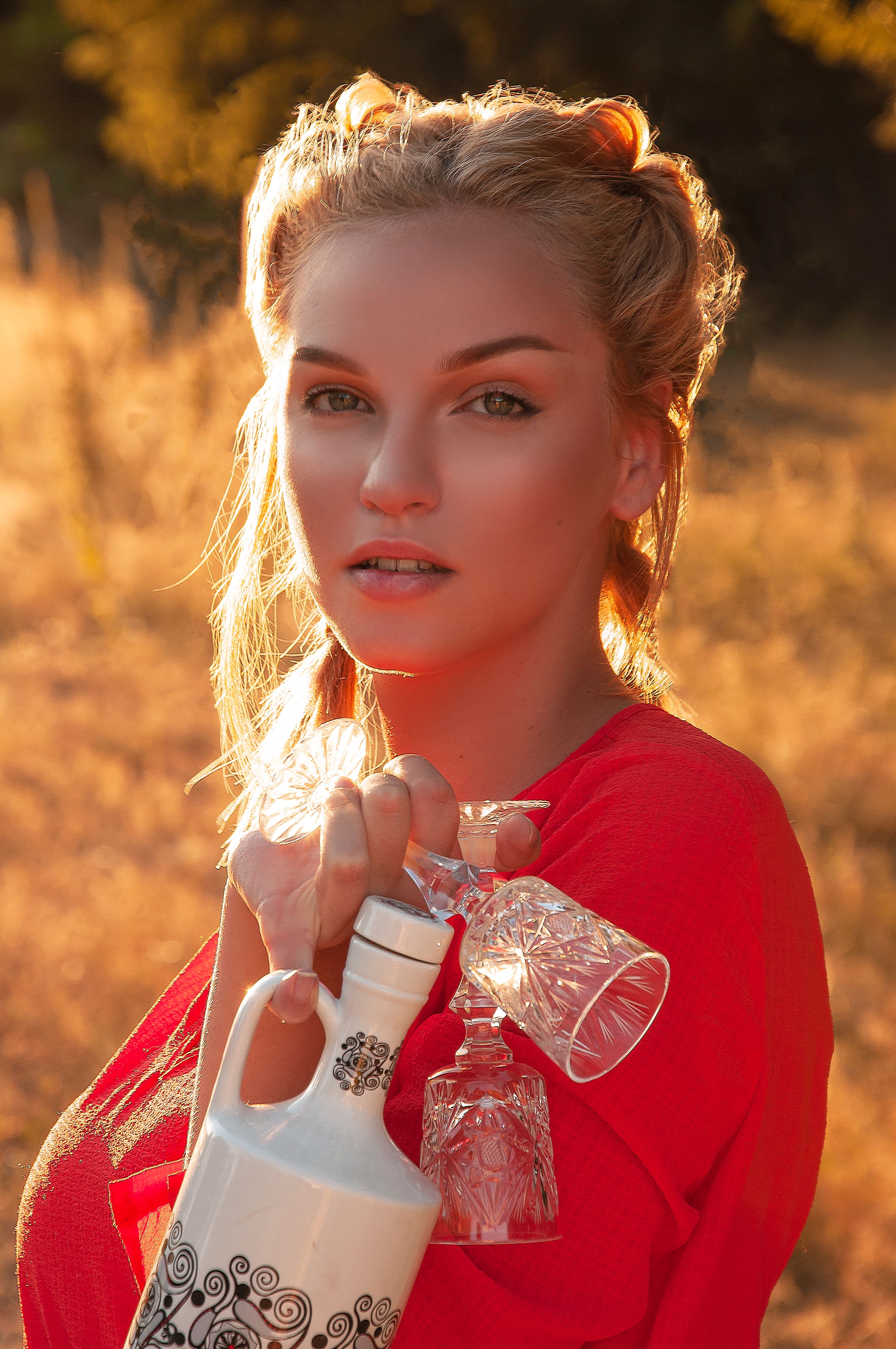 Charming young female with makeup standing with ceramic bottle and glasses in grassy meadow and looking at camera