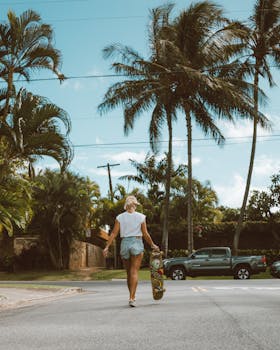 A young woman is walking along a tropical street holding a skateboard, surrounded by palm trees on a sunny day.