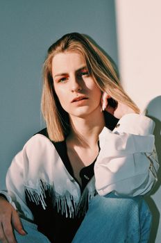 Pensive young lady in stylish outfit touching blond hair while sitting near white wall in sunlight