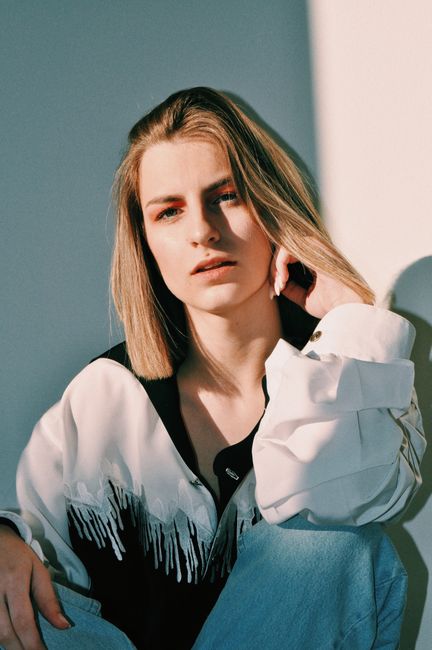 Pensive young lady in stylish outfit touching blond hair while sitting near white wall in sunlight