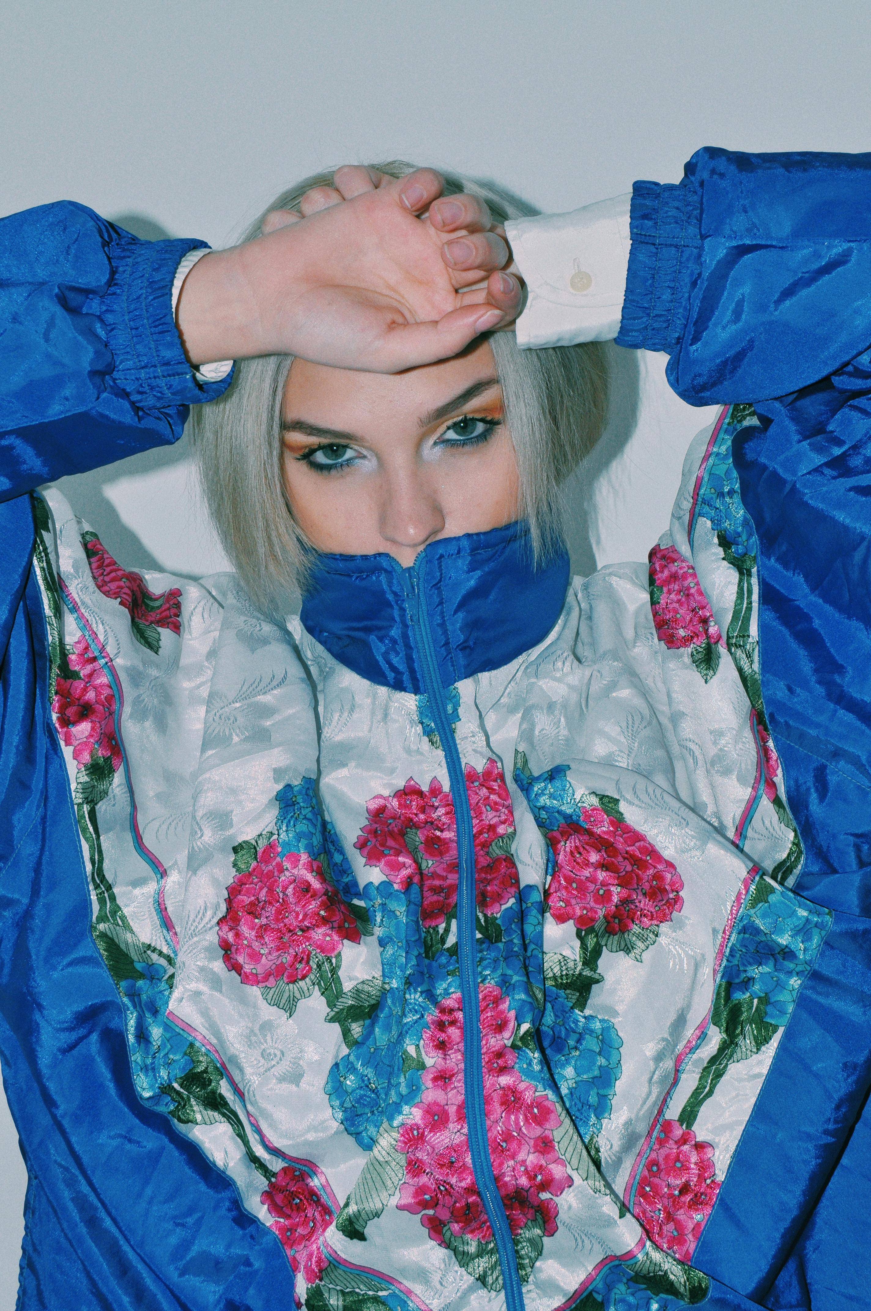 Rebellious young female model with dyed hair covering mouth with collar of trendy jacket and looking at camera