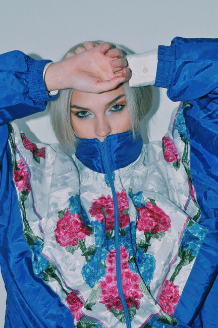 Rebellious young female model with dyed hair covering mouth with collar of trendy jacket and looking at camera