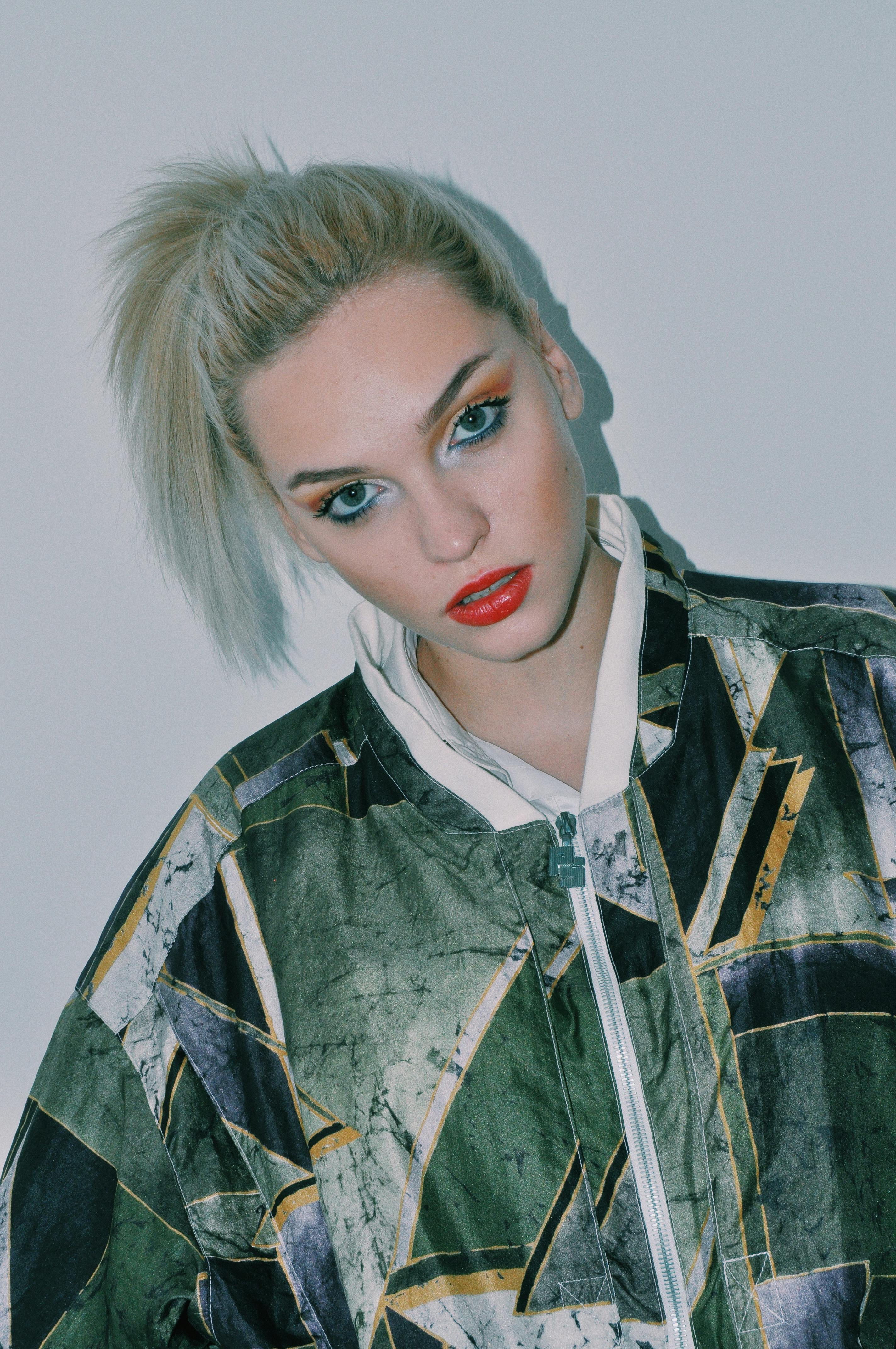 Confident young female model with dyed hair and bright makeup in trendy jacket standing against white background and looking at camera
