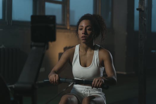 Determined woman in white workout attire rowing indoors, highlighting fitness and focus.