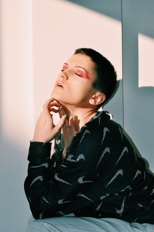 Side view of contemplative young female with short hair and bright eyeshadow touching neck with closed eyes while sitting near white wall in sunlight