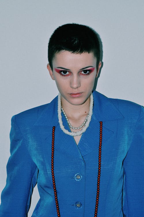 Serious young woman with short dark hair and makeup in elegant jacket standing against white wall and looking at camera