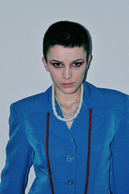 Serious young woman with short dark hair and makeup in elegant jacket standing against white wall and looking at camera