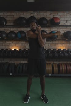 Muscular man stretches in a gym setting surrounded by weights and medicine balls.