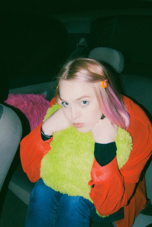 From above of content female teenager with dyed hair in casual clothes embracing soft pillow and looking at camera while sitting in car at night