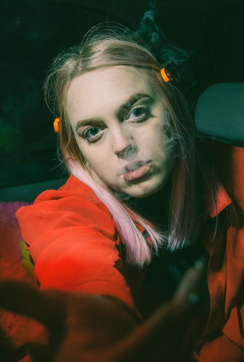 Melancholic young lady with blond hair in casual clothes reaching out hand toward camera while smoking in car at night