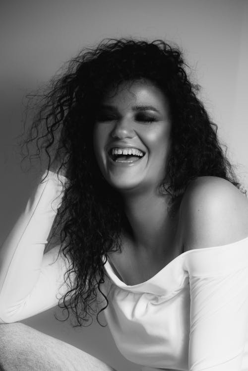 Black and white of cheerful young ethnic female with bare shoulders makeup laughing and touching long curly hair with closed eyes