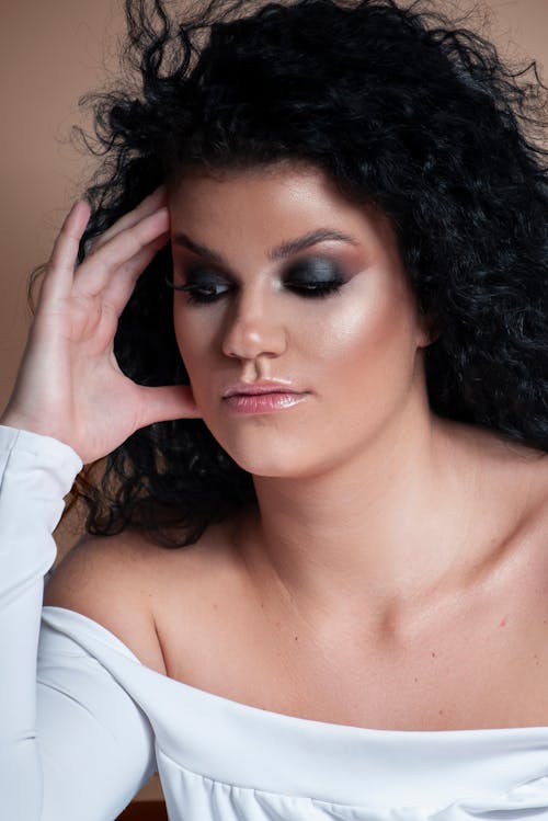 Calm young gorgeous ethnic female model with perfect makeup and long curly hair touching face and looking down against beige background