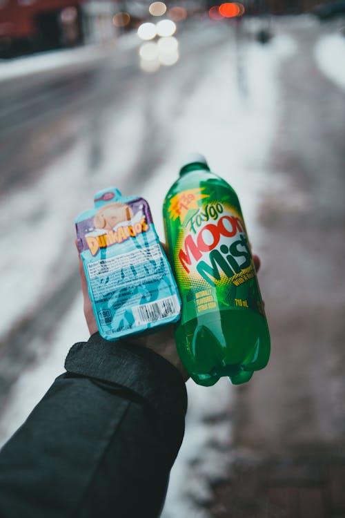 Crop anonymous person showing bottle of carbonated drink and pack of biscuits in winter town