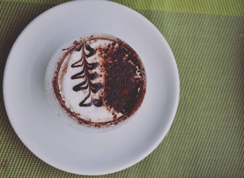 A beautifully crafted cappuccino with artistic cocoa design on a white plate, perfect for a sweet treat.