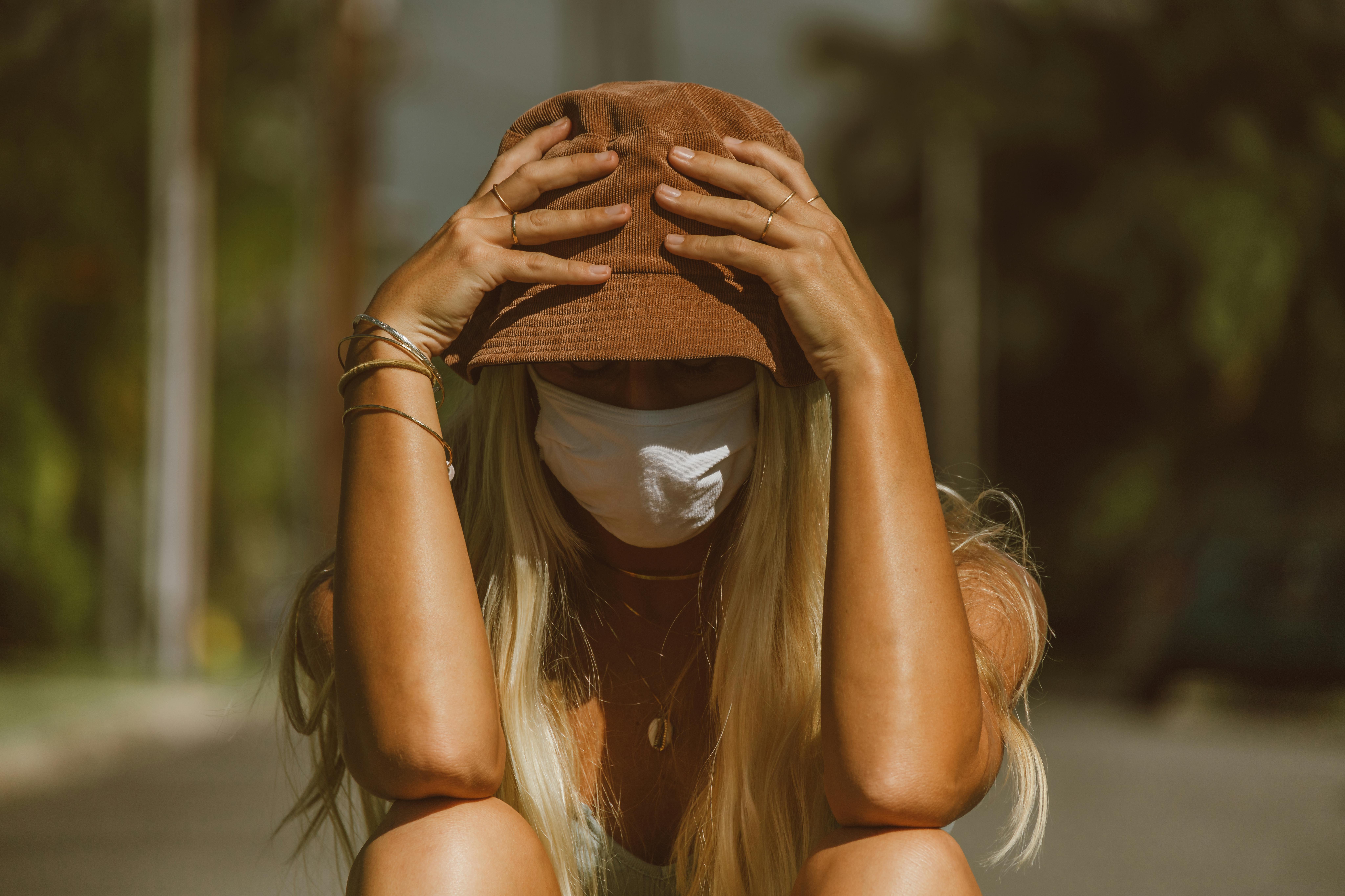 A woman wearing a hat and face mask sits outdoors, embodying a relaxed summer vibe.