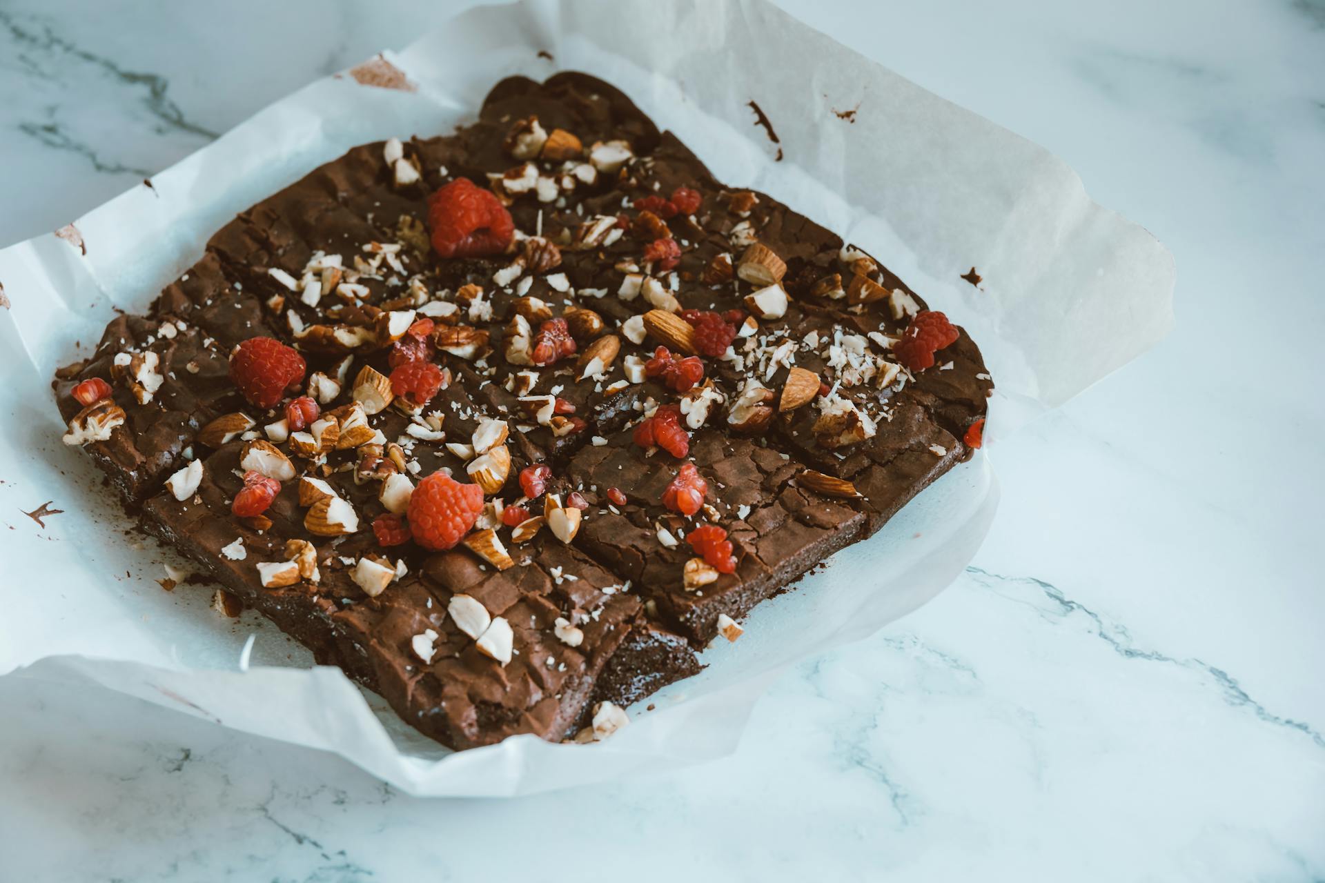 Freshly baked chocolate brownie topped with almonds and raspberries on parchment paper.