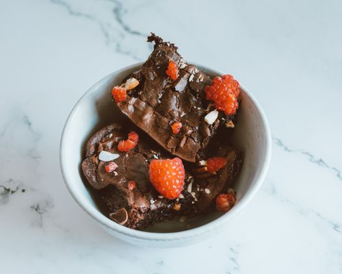 Free A bowl filled with rich chocolate brownies topped with fresh raspberries and nuts on a white marble surface. Stock Photo